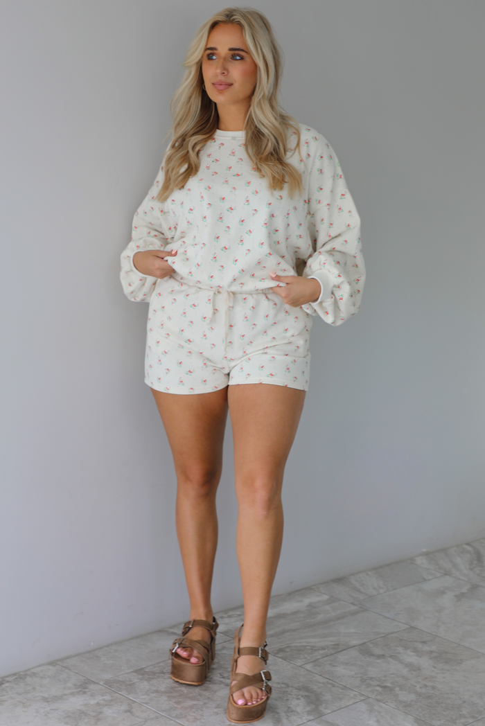 Woman wearing a two piece cream set that has a floral design throughout featuring a sweatshirt and sweat-shorts indoors against a white wall and white marble flooring. 