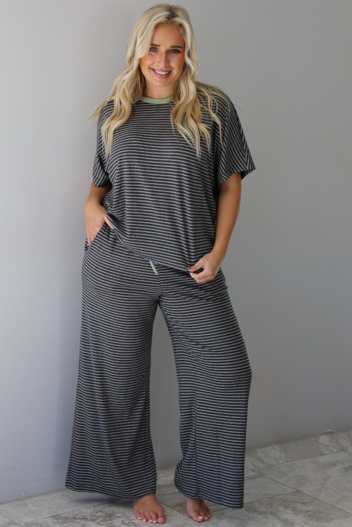 A woman wearing a charcoal and white striped two piece matching set with green trim detailing at the neckline and waistline, featuring an oversized short sleeved top and high waisted pants, standing indoors against a white wall with white marble flooring. 