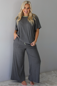 A woman wearing a charcoal and white striped two piece matching set with green trim detailing at the neckline and waistline, featuring an oversized short sleeved top and high waisted pants, standing indoors against a white wall with white marble flooring. 