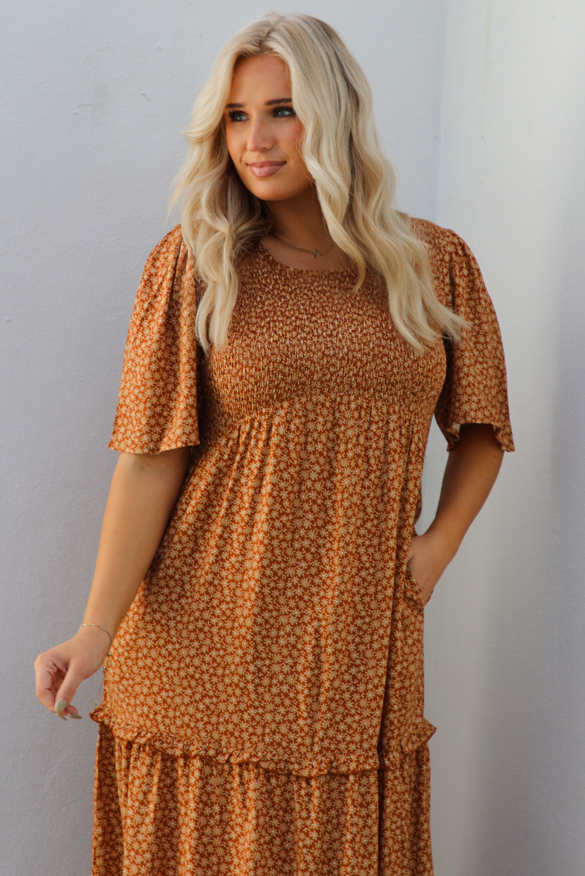 Up close of a woman model outdoors wearing a rust dress with a cream floral print pattern throughout. It has flowy short sleeves, a smocked bodice, and a flowy maxi length skirt with functional pockets on either side. 