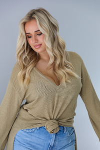 Woman wearing a camel long-sleeve top that's tied in the front with a knot and has a train in the back. The photo shows a closeup of the top which has small holes in it! The photo is taken indoors against a white wall.