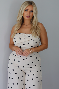 A woman wearing a sand strapless jumpsuit with a black polka dot print pattern throughout, standing indoors against a white wall. The photo is focused on the bodice of the jumpsuit.