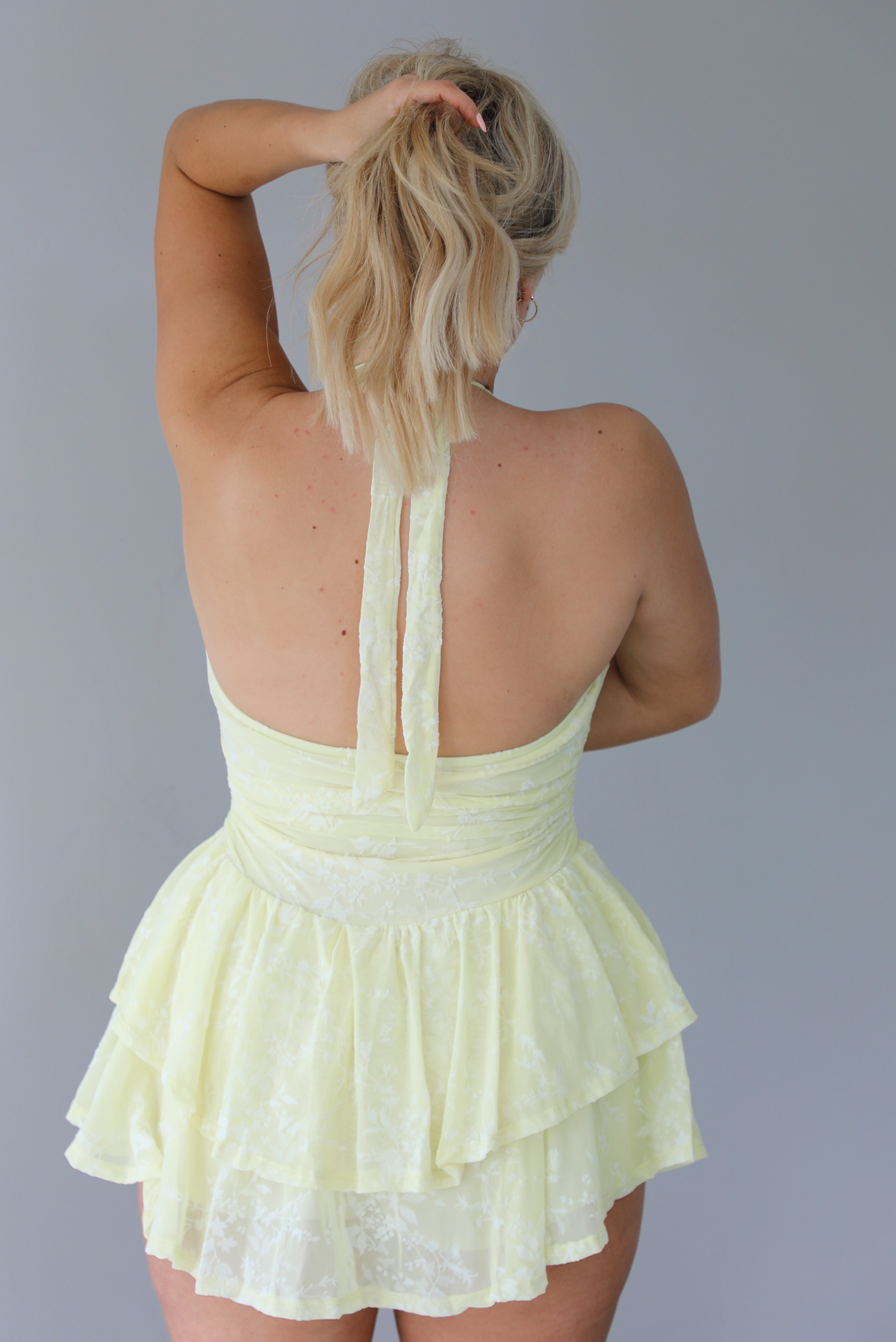 Woman wearing a pale yellow romper that looks like a dress that has a white embroidered floral design, a halter tie neckline, and a ruffled skirt. The photo is taken indoors against a white wall and shows the back of the romper. 