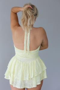 Woman wearing a pale yellow romper that looks like a dress that has a white embroidered floral design, a halter tie neckline, and a ruffled skirt. The photo is taken indoors against a white wall and shows the back of the romper. 