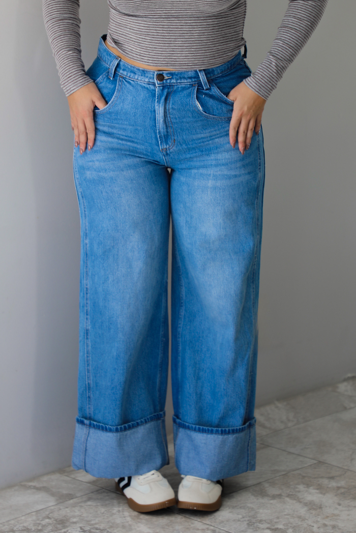 A closeup of a woman wearing high waisted medium wash denim jeans with a cuffed hem, standing indoors against a white wall with white marble flooring. 
