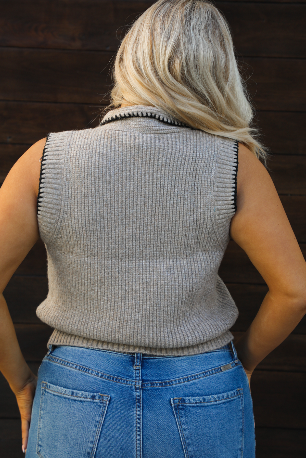 Back of a woman model wearing a taupe sweater vest with black stitched detailing, has a tie detail at the neckline, is sleeveless, has functional button closures in the front, and has a ribbed texture. The sweater vest is paired with a medium wash denim. The photo is taken outdoors against a dark brown wooden door. 
