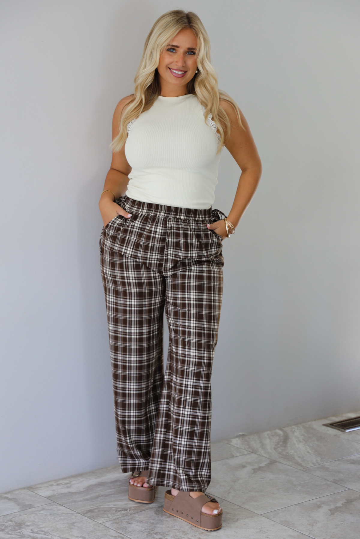 A woman modeling a pair of brown and sand plaid wide-leg pants with a cream ribbed sleeveless top, standing indoors against a white wall with white marble flooring. 