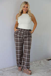A woman modeling a pair of brown and sand plaid wide-leg pants with a cream ribbed sleeveless top, standing indoors against a white wall with white marble flooring. 