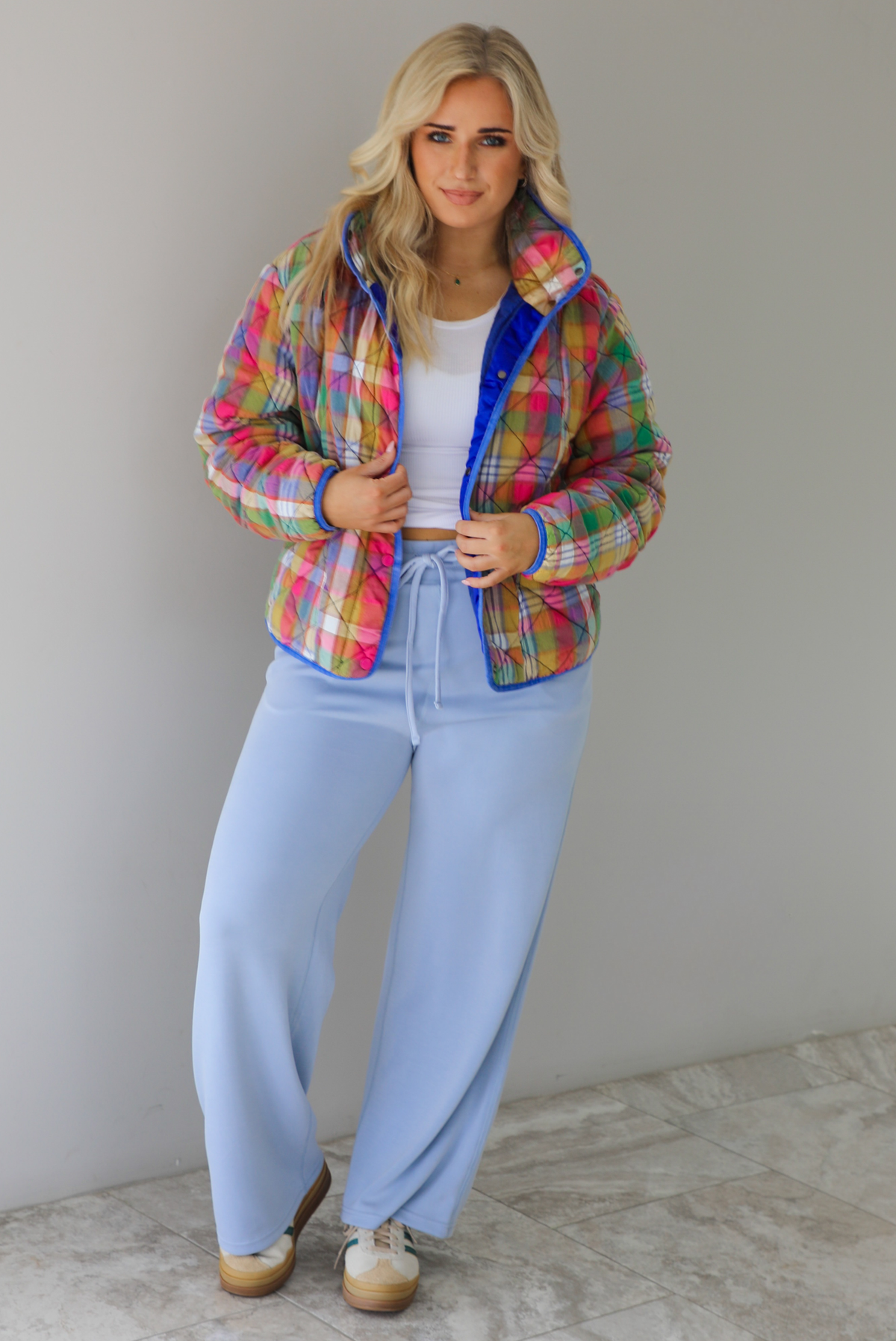 Woman wearing a multi-colored plaid, quilted style jacket paired with a white top underneath and high waisted blue lounge pants. The photo is taken indoors against a white wall with white marble flooring. 