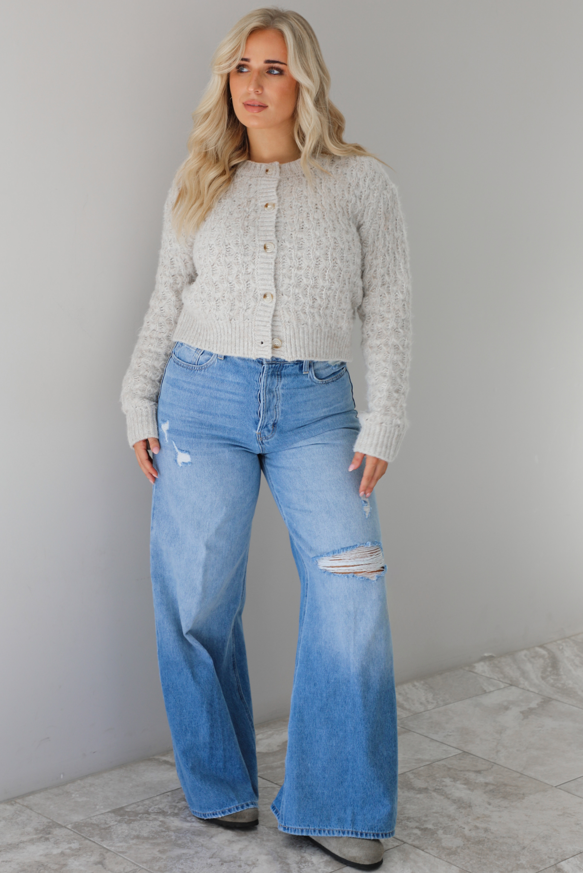 Woman wearing a light grey cardigan buttoned up with high waisted flared hem medium wash denim. The photo is taken indoors against a white wall with white marble flooring.