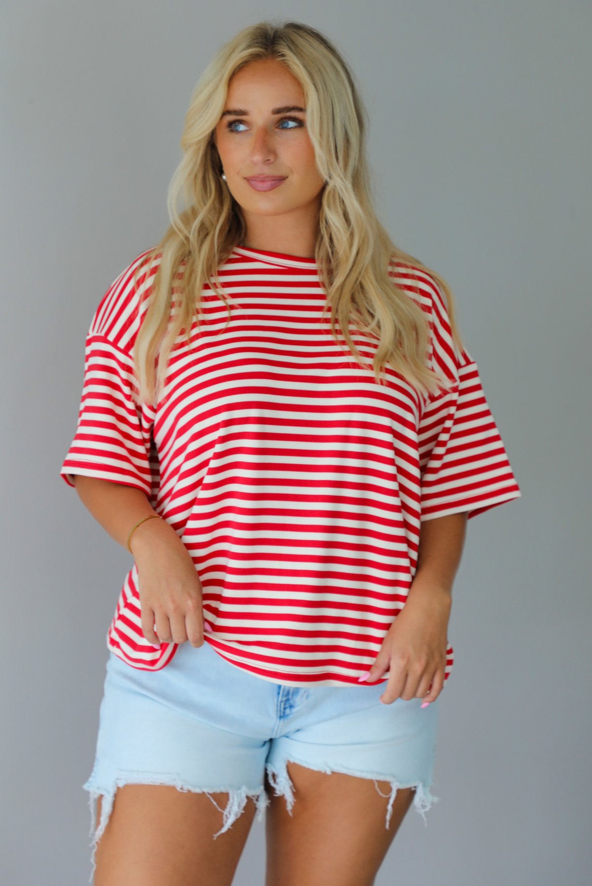 A woman modeling a red and white horizontally striped short-sleeve t-shirt with light blue denim shorts and brown sandals, standing indoors against a white wall. The top is untucked from the shorts to show the full length. 
