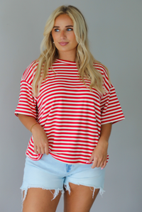 A woman modeling a red and white horizontally striped short-sleeve t-shirt with light blue denim shorts and brown sandals, standing indoors against a white wall. The top is untucked from the shorts to show the full length. 
