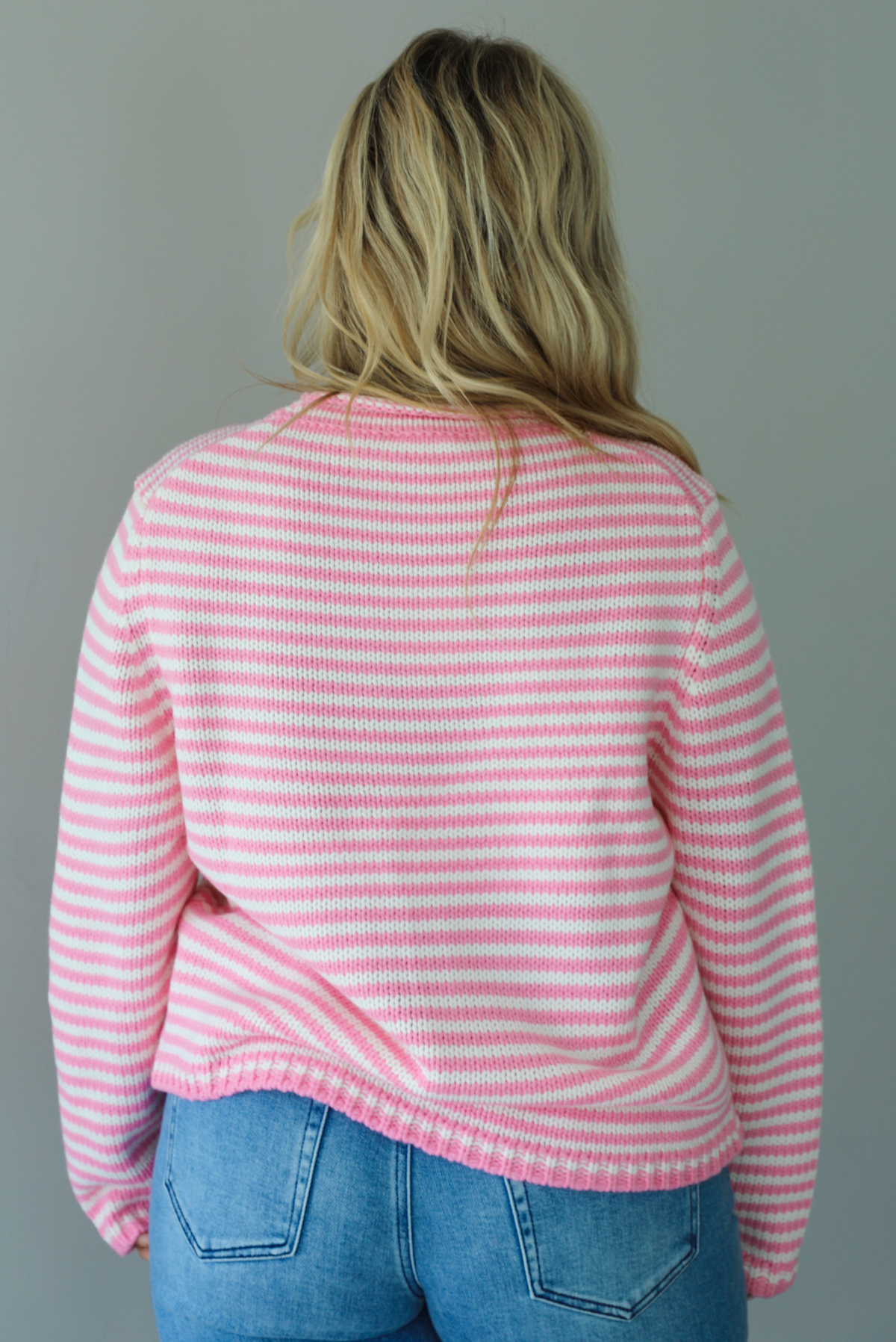 A woman shown from behind wearing a pink and white striped sweater paired with light wash denim jeans, standing indoors against a white wall.