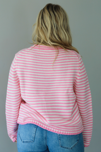 A woman shown from behind wearing a pink and white striped sweater paired with light wash denim jeans, standing indoors against a white wall.