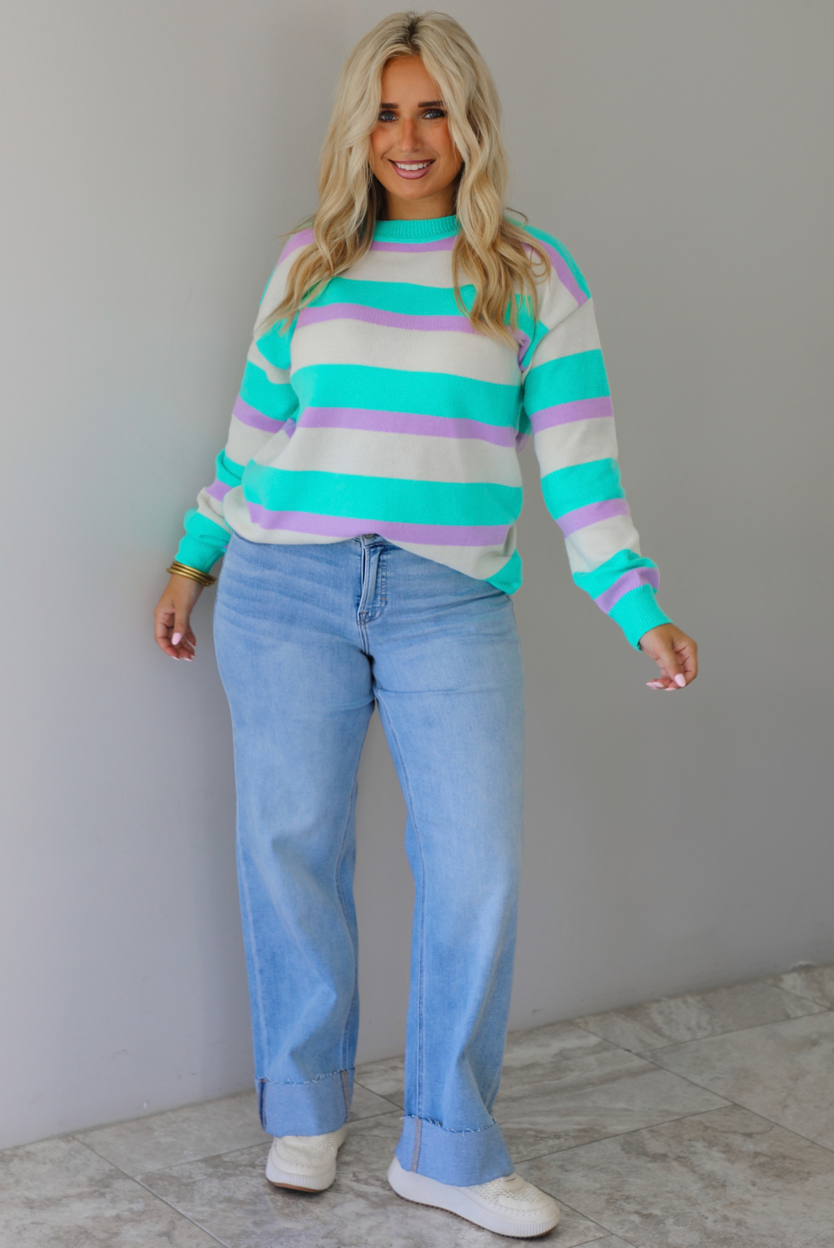 A woman wearing a turquoise, lavender, and cream striped knit sweater, paired with light wash denim jeans, standing indoors against a white wall with white marble flooring. 