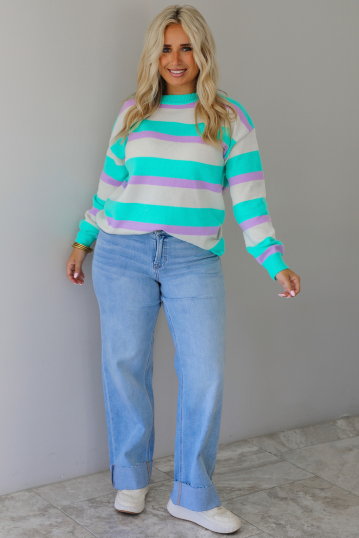 A woman wearing a turquoise, lavender, and cream striped knit sweater, paired with light wash denim jeans, standing indoors against a white wall with white marble flooring. 