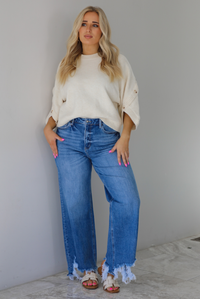 Woman wearing a cream colored quarter sleeve sweater that has ribbed details at the neckline, on the sleeves, and on the hem, with non-functional button closures on the sleeves. The sweater has a relaxed fit and is paired with a medium wash, high waisted, distressed hem denim with sand colored sandals. The photo is taken indoors against a white wall with white marble flooring. 
