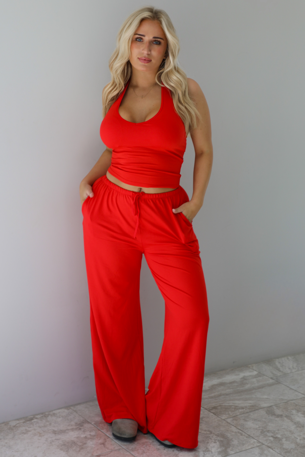 Woman wearing a red two piece set featuring a  tank top and slouchy pants. The photo is taken indoors against a white wall with white marble flooring. 