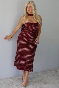 A woman wearing a strapless maroon midi dress with a white polka dot pattern with a matching scarf, standing indoors against a white wall with white marble flooring. 