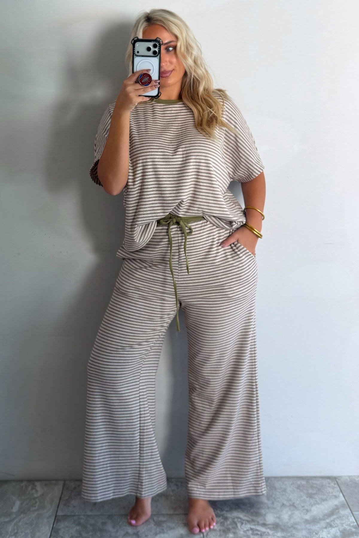 A woman takes a mirror selfie wearing a two-piece loungewear set consisting of a short-sleeve top and matching wide-leg pants with thin horizontal stripes in beige and olive green, featuring an olive green drawstring waist.