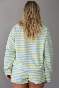 Woman wearing a blue and pale yellow striped two piece set featuring an oversized sweatshirt and shorts. The photo is taken indoors against a white wall and shows a closeup of the top of the set, which is untucked to show the full length of the top. 