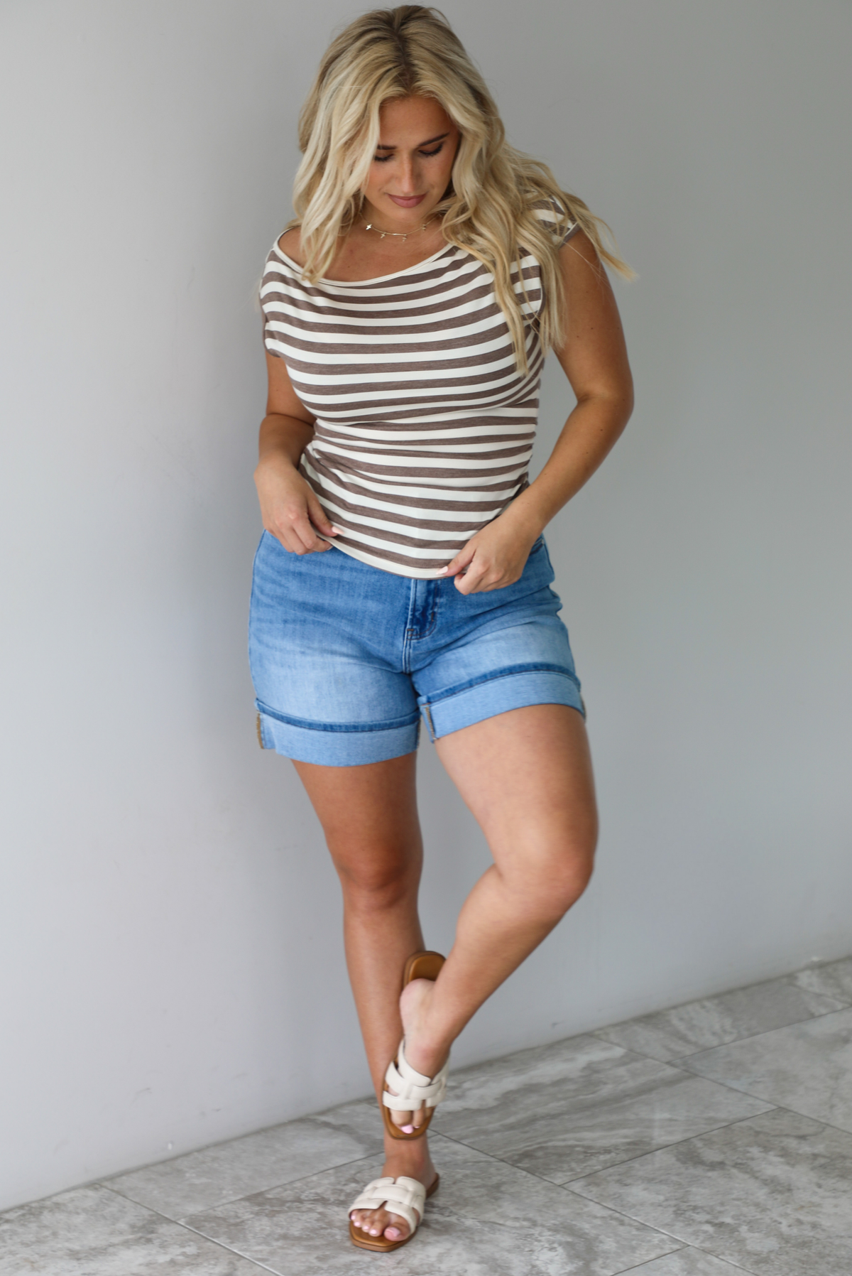 Woman wearing a mocha and white striped top with an off the shoulder design with medium wash denim shorts indoors against a white wall with white marble flooring. 