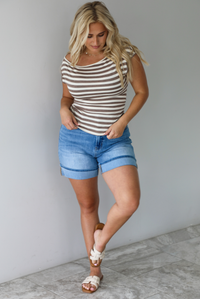 Woman wearing a mocha and white striped top with an off the shoulder design with medium wash denim shorts indoors against a white wall with white marble flooring. 