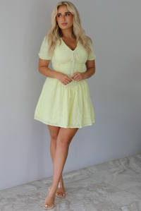 Woman wearing a light yellow mini dress with a floral eyelet design throughout, v-neckline, short sleeves, fitted drop waist bodice, and a flowy mini length skirt indoors against a white wall with white marble flooring. 
