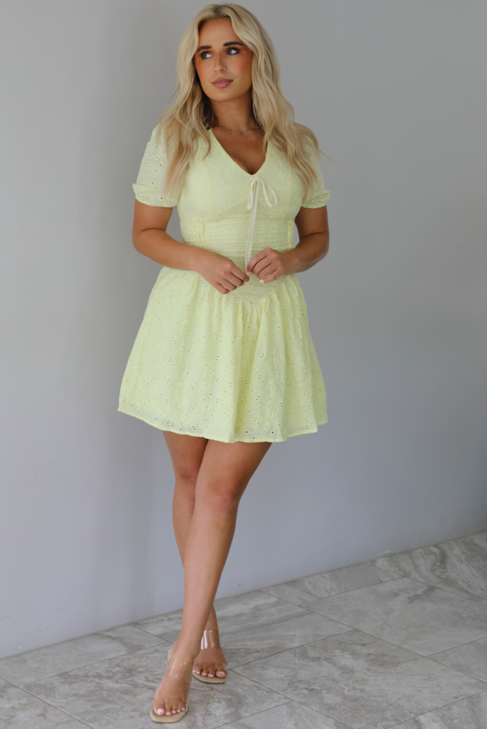 Woman wearing a light yellow mini dress with a floral eyelet design throughout, v-neckline, short sleeves, fitted drop waist bodice, and a flowy mini length skirt indoors against a white wall with white marble flooring. 