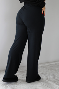 The photo features a model wearing high waisted black pants that have an elastic and drawstring waistline, a slightly flared hem, and are super soft and stretchy. They are worn with black ugg boots. The photo is taken indoors against a white wall with white marbled flooring. The photo shows the back of the pants. 