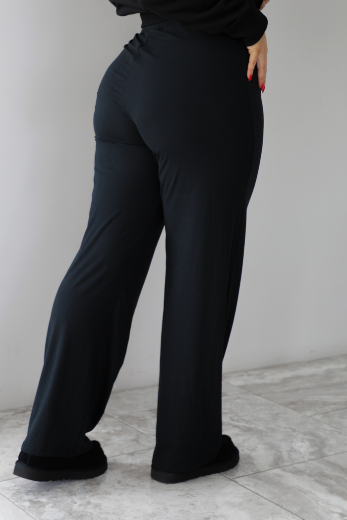 The photo features a model wearing high waisted black pants that have an elastic and drawstring waistline, a slightly flared hem, and are super soft and stretchy. They are worn with black ugg boots. The photo is taken indoors against a white wall with white marbled flooring. The photo shows the back of the pants. 