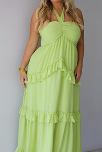 A woman modeling a lime green halter-neck maxi dress with tiered ruffle details, standing indoors against a white wall.