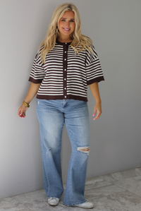 A woman wearing a brown and white striped knit button top cardigan top, paired with light wash denim distressed denim, standing indoors against a white wall with white marble flooring. 