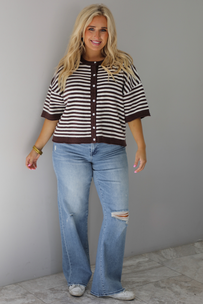 A woman wearing a brown and white striped knit button top cardigan top, paired with light wash denim distressed denim, standing indoors against a white wall with white marble flooring. 