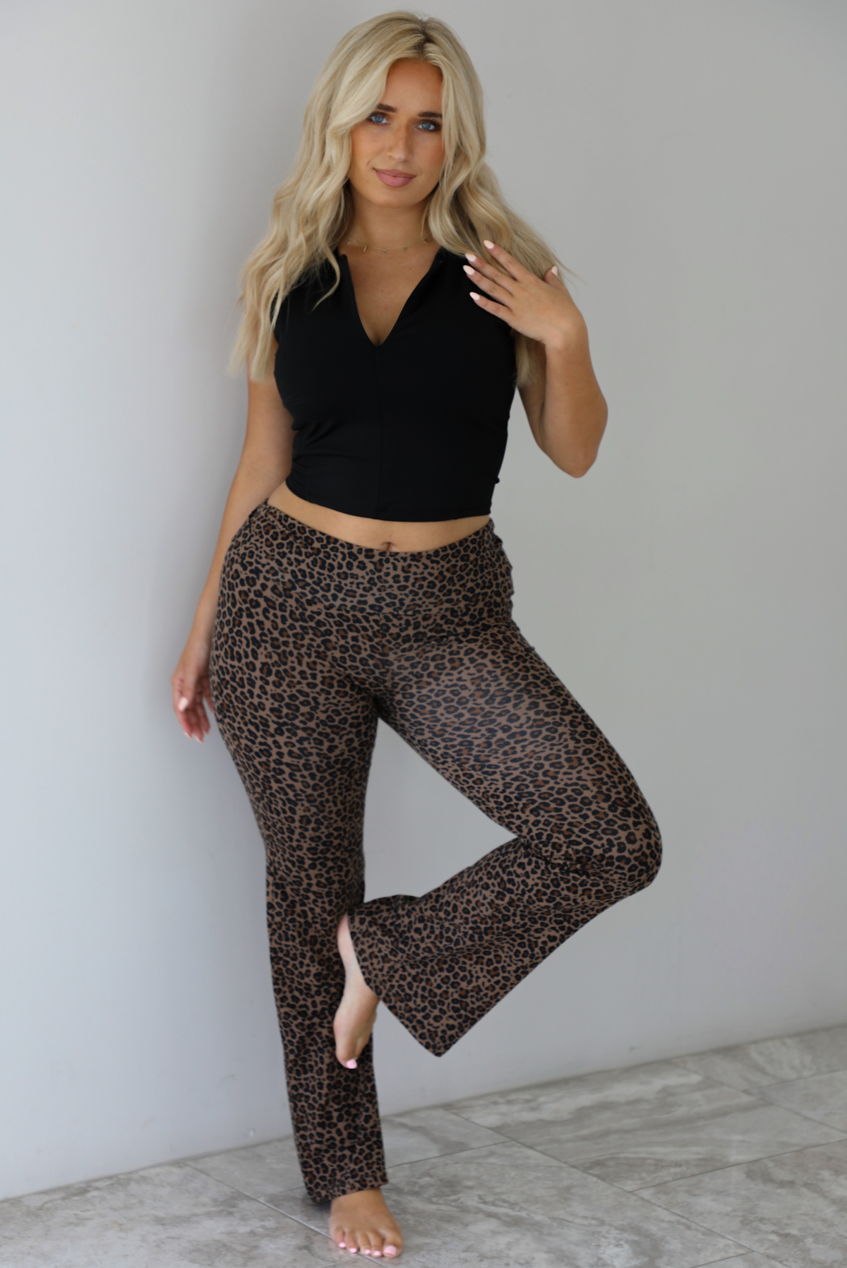 Woman wearing a black quarter zip sleeveless crop top with mid-rise leopard casual pants indoors against a white wall with white marble flooring. 