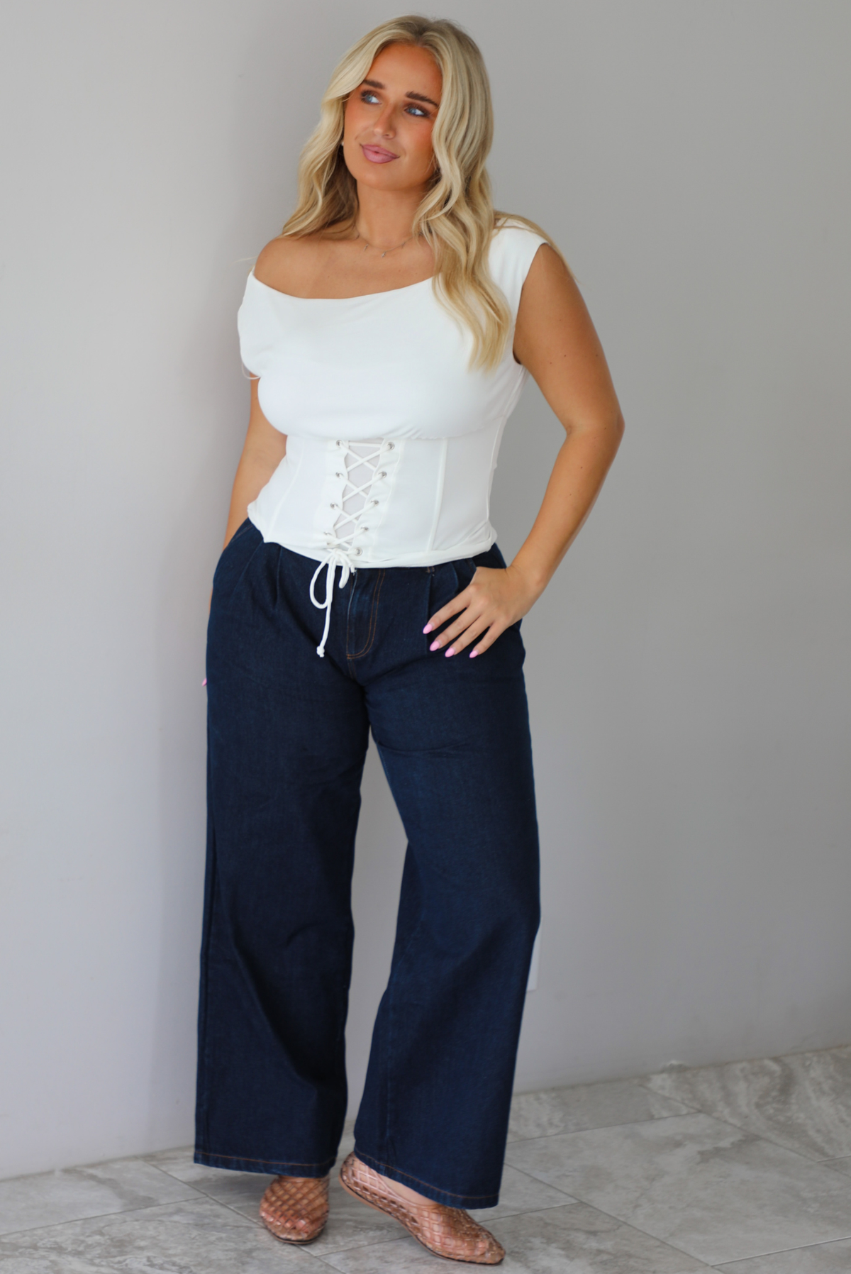 A woman modeling a white off-the-shoulder lace-up corset top paired with dark denim wide-leg jeans and pinkish-brown flat shoes, standing indoors against a white wall with white marble flooring. 