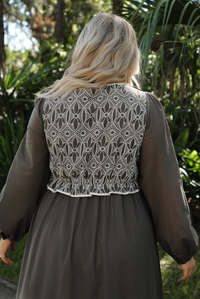 Close up of the back of a woman model wearing an olive and off-white stitched detail dress outdoors. Dress has a corset-appearance bodice, long sleeves, functional pockets on either side, an elastic waistline, and a long, maxi length.