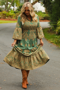 Woman wearing a teal dress that has tan a stitched pattern throughout, quarter length  sleeves, a collared, v-neckline, cinched with elastic waistline, and a maxi-length skirt. The photo is taken outdoors standing on a paved road with trees and other greenery in the background.