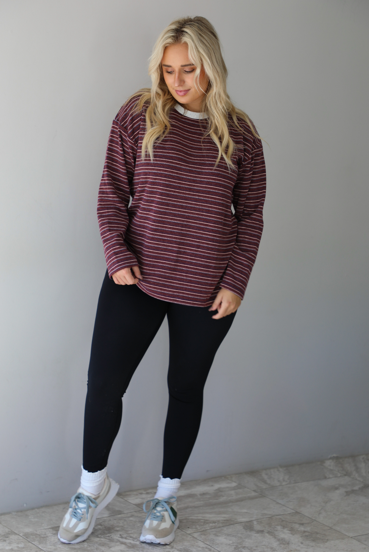 A woman wearing a burgundy striped long sleeved top paired with black leggings, high ankle socks, and sneakers, standing indoors against a white wall with white marble flooring. 
