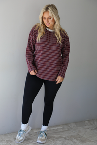 A woman wearing a burgundy striped long sleeved top paired with black leggings, high ankle socks, and sneakers, standing indoors against a white wall with white marble flooring. 