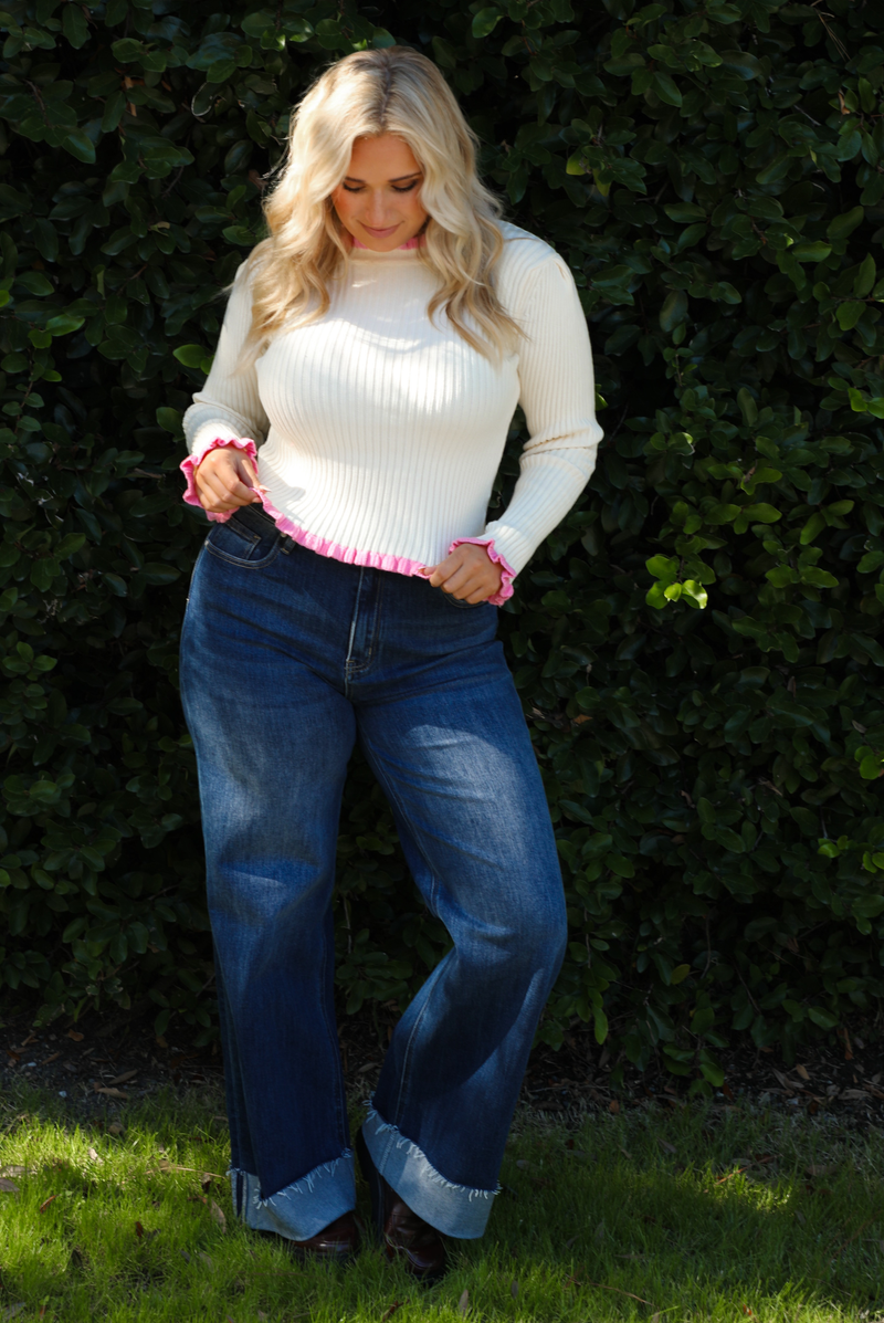 Woman model wearing a cream knit top with pink trim and dark denim jeans. The photo is taken outdoors against greenery. 