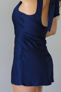 A woman shown from the side, focused on the zipper closure, wearing a navy blue sleeveless dress with a large bow detail at the open back, standing indoors against a white wall.