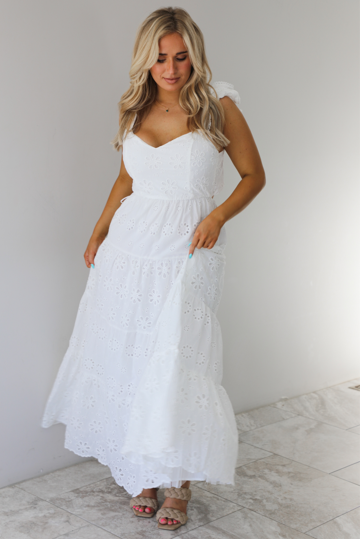 Woman wearing a white maxi length dress that has an eyelet floral design throughout and has a fitted bodice. The photo is taken indoors against a white wall with white marble flooring. 