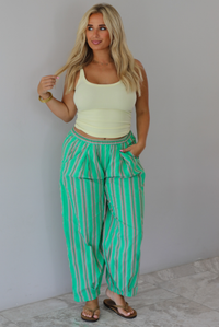 A woman wearing a pale yellow tank and mid-rise pants that have a green, brown, lime green, and tan striped pattern, which have an elastic waistline, functional pockets on either side, and cinched details at the ankles, standing indoors against a white wall with white marble flooring. 
