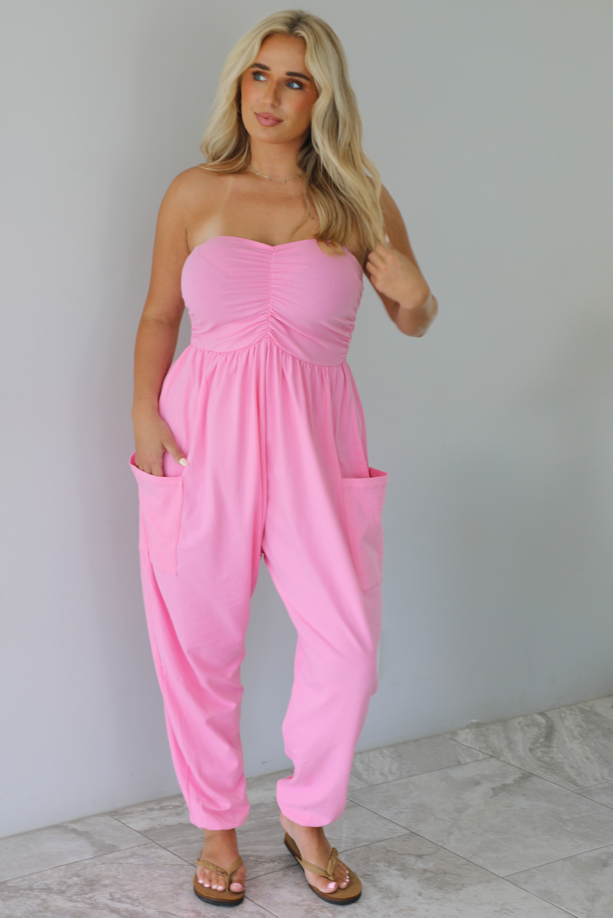 Woman wearing a pink strapless jumpsuit with a fitted bodice and slouchy pants indoors against a white wall with white marble flooring. 