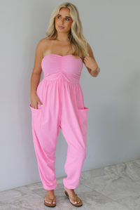 Woman wearing a pink strapless jumpsuit with a fitted bodice and slouchy pants indoors against a white wall with white marble flooring. 