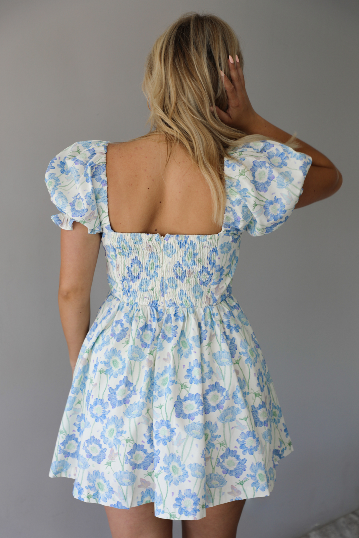 Woman wearing a white, blue, and green floral print mini dress that has short, puffed sleeves, a fitted corset bodice, and a flowy skirt indoors against a white wall and shows the back of the dress, which has a smocked back with a zipper closure. 