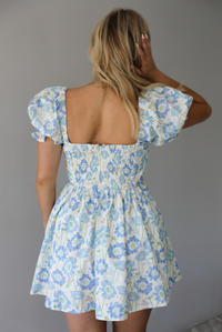 Woman wearing a white, blue, and green floral print mini dress that has short, puffed sleeves, a fitted corset bodice, and a flowy skirt indoors against a white wall and shows the back of the dress, which has a smocked back with a zipper closure. 
