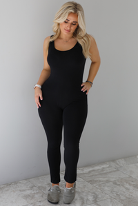 Woman wearing a black fitted jumpsuit with thick tank-top style straps. The jumpsuit is paired with grey slide on shoes. The photo is taken indoors in front of a white wall with white marble flooring. 