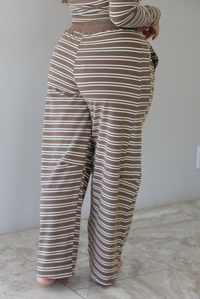 A woman wearing a brown with cream striped matching two piece set consisting of a long sleeved, off the shoulder top and high waisted pants, standing indoors against a white wall with white marble flooring. The photo shows a closeup of the pants back of the pants. 